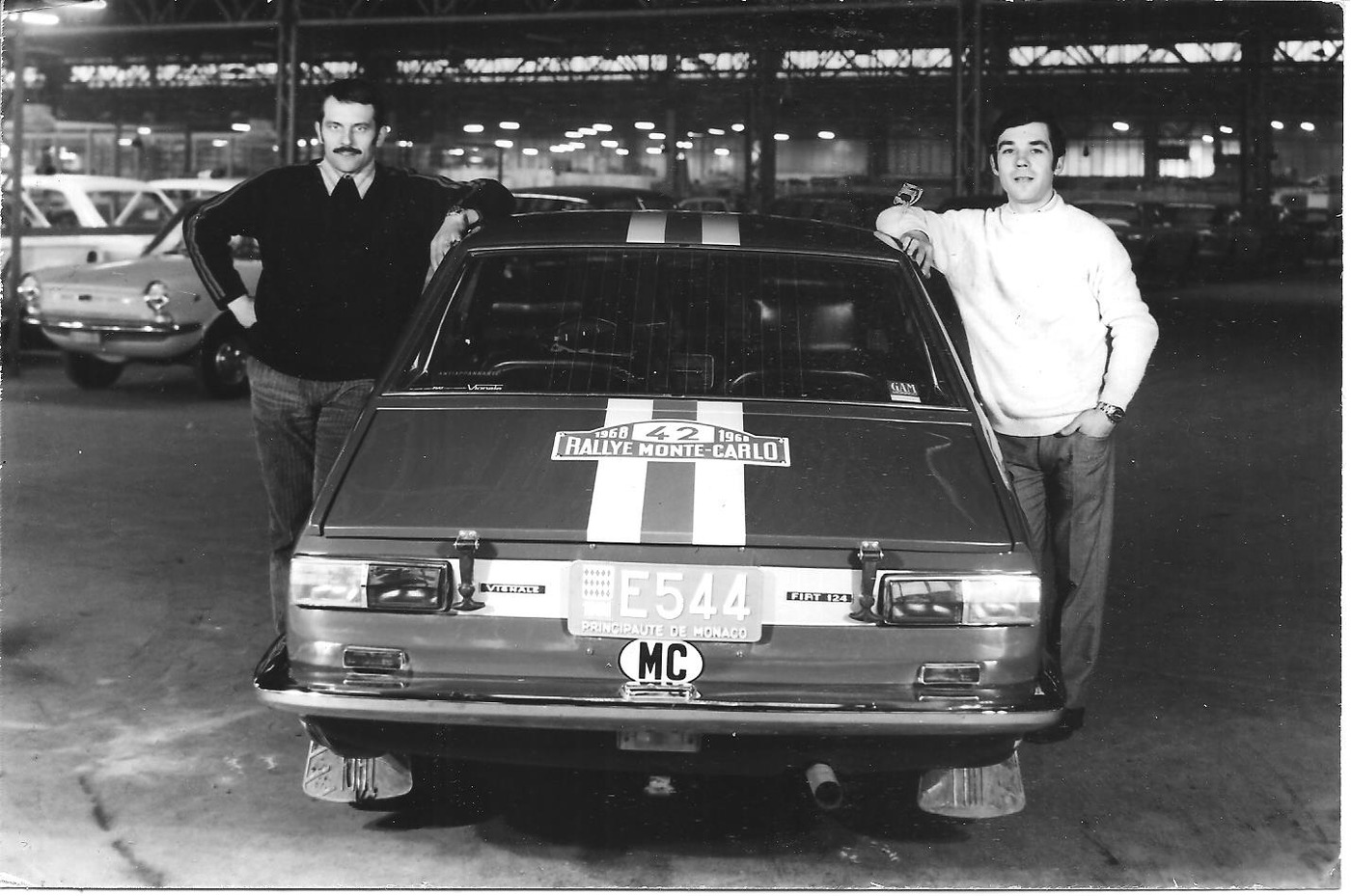 Two crew members standing behind a Fiat 124 Vignale with racing stripes, a GAM sticker and a Rallye Monte-Carlo rear-window plaque, Monaco plate E544